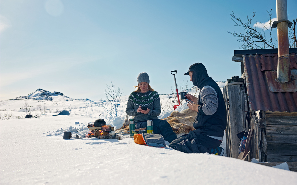 En kvinna och man på picknick i ett vinterlandskap med termosar framför sig, sittandes vid en öppen eld och kvinnan surfar på sin mobil.